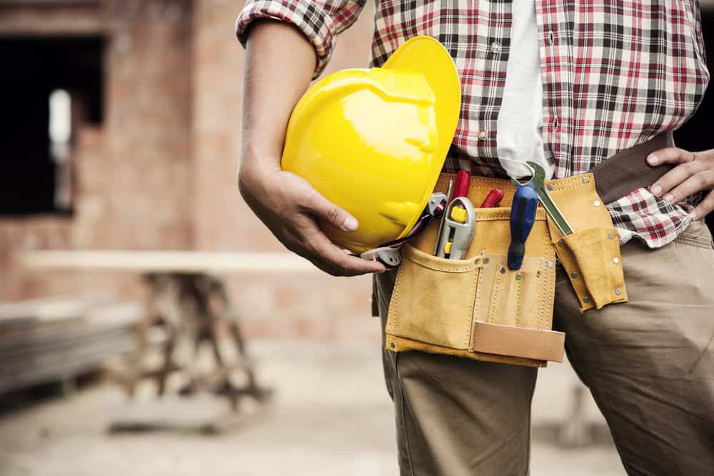 A construction worker wearing a plaid shirt and tool belt holds a yellow hard hat. The tool belt contains various tools, and the background shows a blurred construction site.