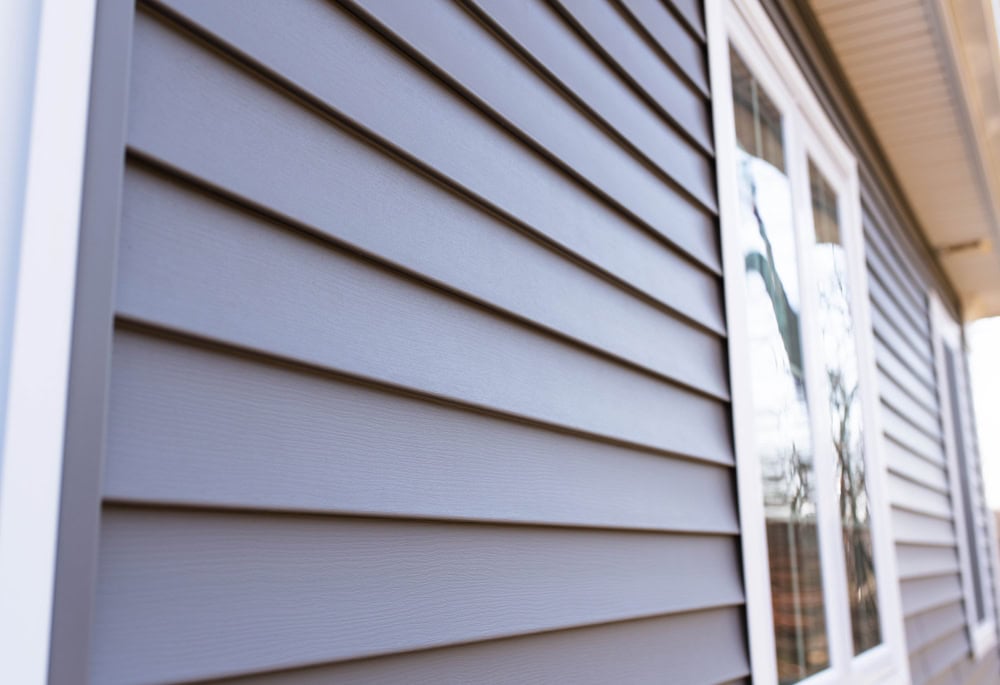 Close-up view of a house exterior showcasing blue-gray horizontal vinyl siding. A portion of a window is visible on the right. The image highlights the smooth, even lines and texture of the siding.