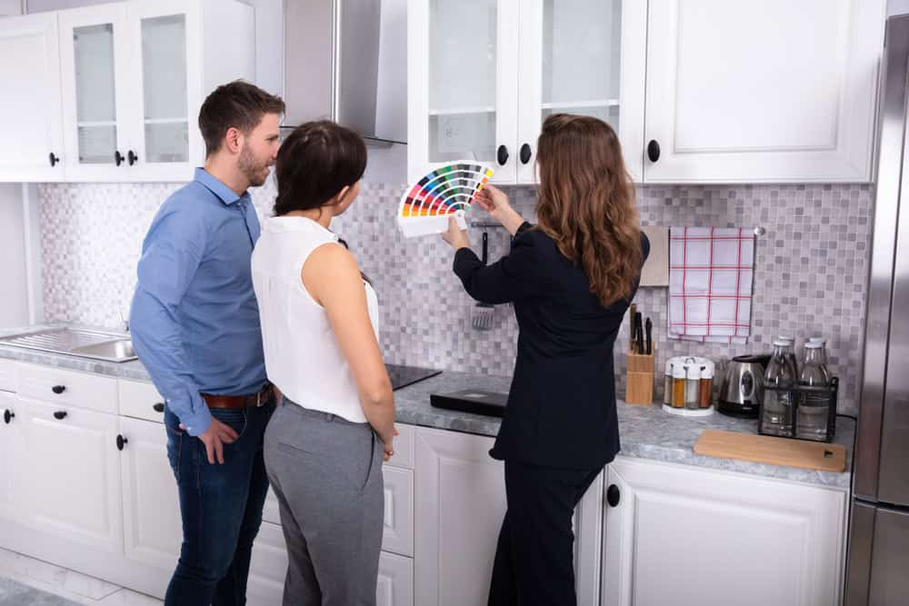 A couple in a kitchen with white cabinets is consulting a designer who is holding a color swatch. They are discussing color options for the kitchen, with various bottles and utensils on the counter in the background.