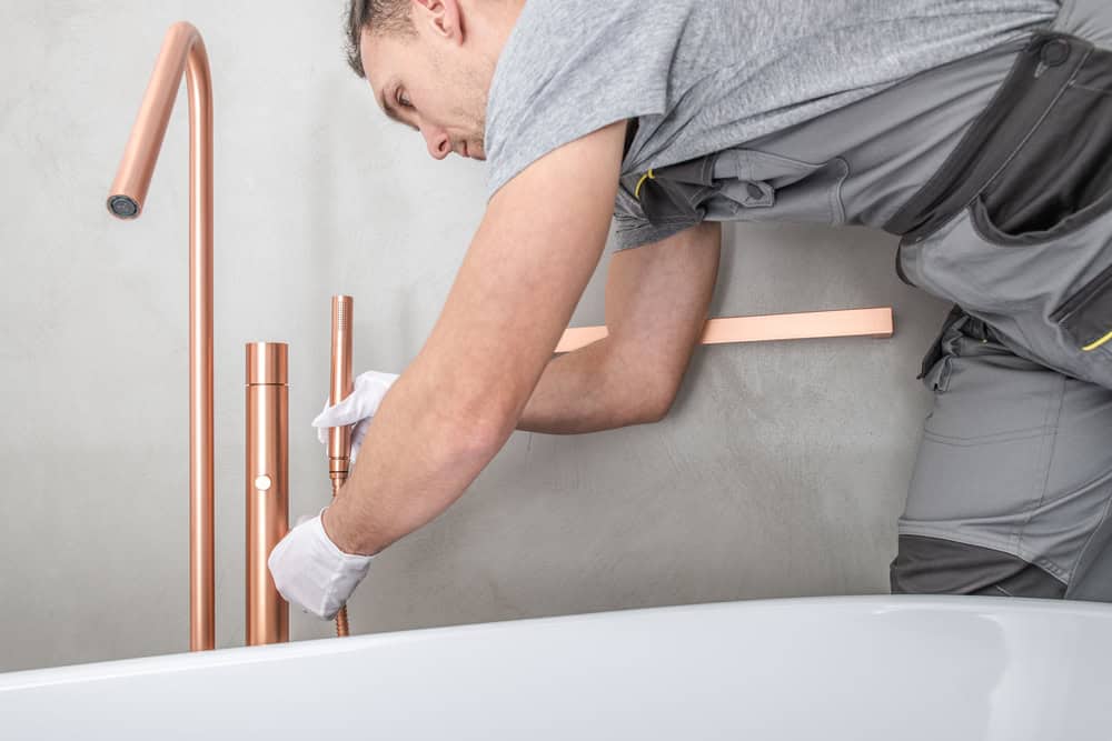 A person in workwear and gloves is installing or repairing a copper-colored faucet next to a bathtub against a light-colored wall.