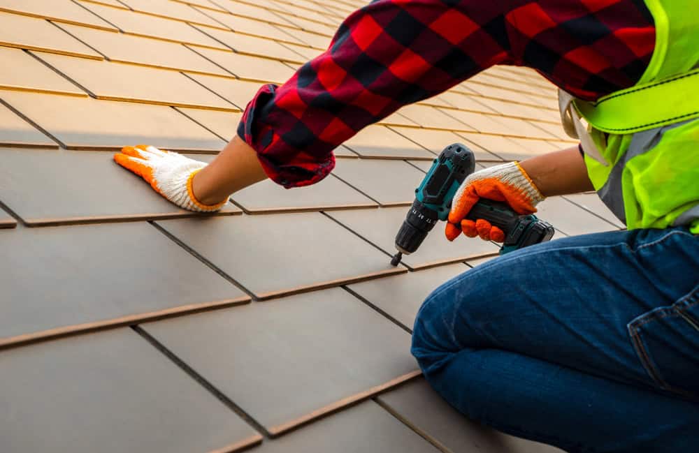 A worker wearing a red plaid shirt, safety vest, and gloves uses a power drill to secure roof tiles. The person is kneeling on a sloped rooftop, focusing on the alignment of the rectangular tiles.