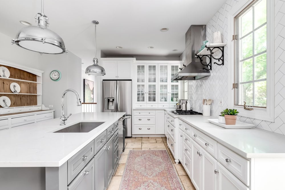 Bright, modern kitchen featuring white cabinetry, stainless steel appliances, and a large island with a farmhouse sink. Two metallic pendant lights hang above. A window lets in natural light, and a potted plant sits on the counter. A rug adds color.