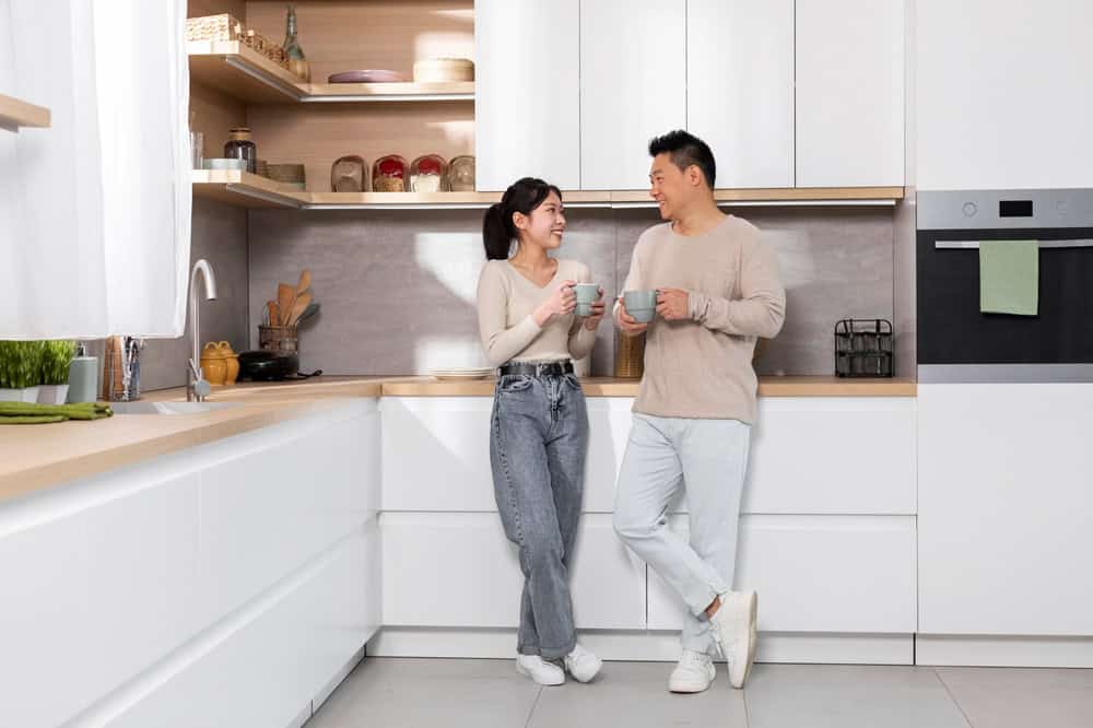 A couple stands in a modern kitchen, holding mugs and smiling at each other. The kitchen has white cabinets, wooden countertops, and open shelves with various items. They both wear casual outfits, complementing the bright, airy space.