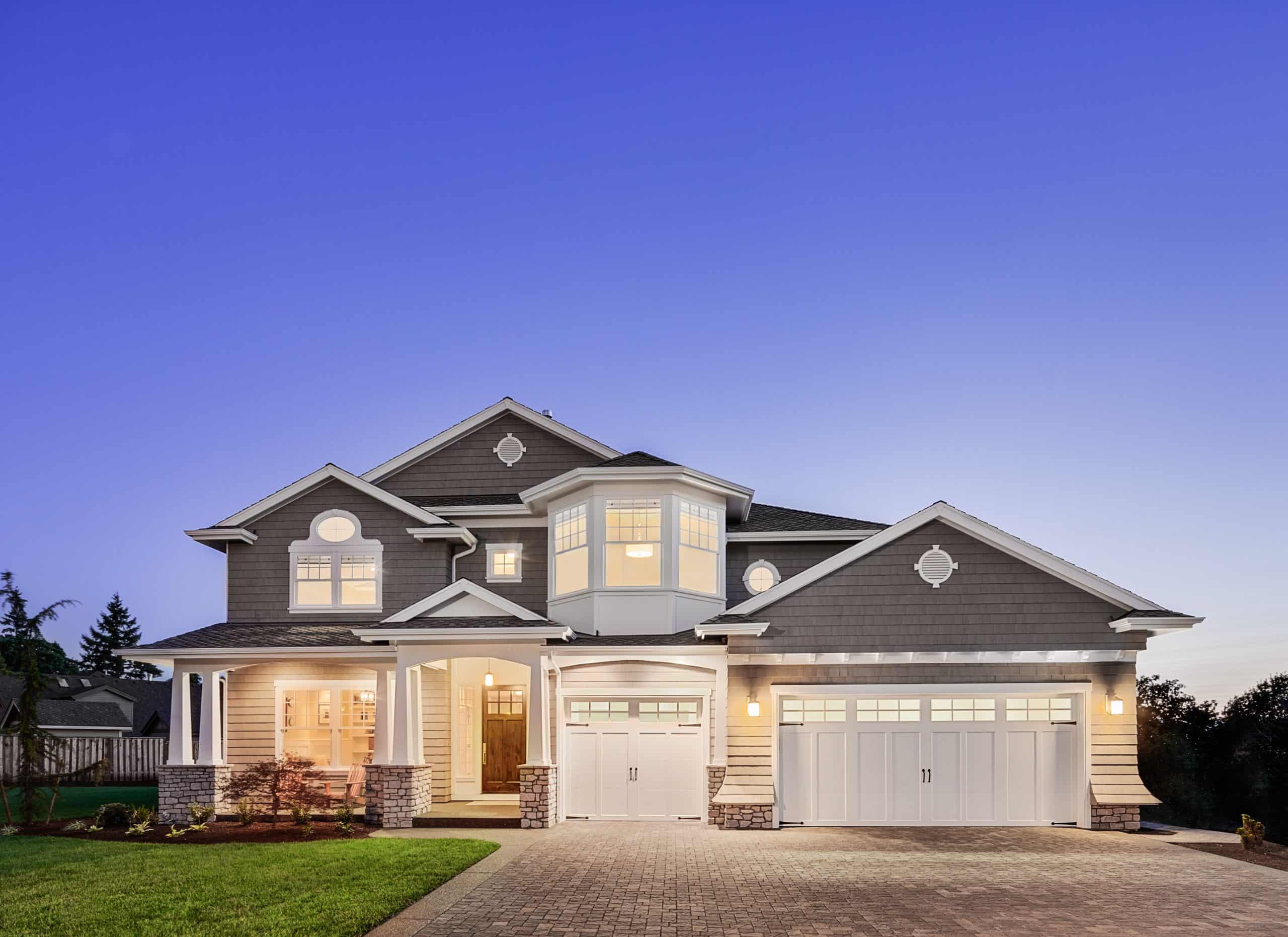 A two-story house with a gray facade and white trim, featuring multiple peaked roofs, a large central bay window, and a double garage. The exterior is well-lit, and the surrounding yard is neatly landscaped with grass and small shrubs.
