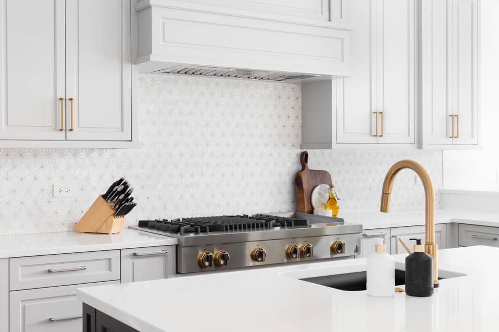 A modern kitchen with white cabinetry and a gas stove featuring gold knobs. White countertops hold a knife block, a soap dispenser, and a cutting board with oil. A gold faucet is above a black sink, and the backsplash has a geometric tile pattern.