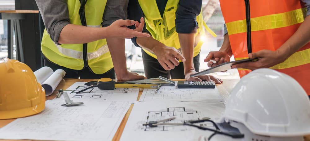A group of people in safety vests discussing building plans on a table. The table is covered with blueprints, a calculator, a ruler, and safety helmets. The focus is on their hands and the documents.