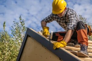 A construction worker wearing a yellow hard hat and gloves is nailing a metal trim on a roof edge. He is kneeling on a wooden structure, with blue sky and trees in the background.