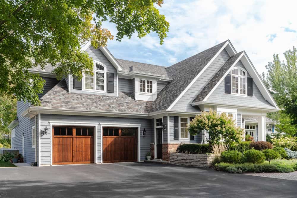 A two-story suburban house with a gray exterior, large windows, and a driveway leading to two wooden garage doors. The yard features lush greenery and trees under a partially cloudy sky.