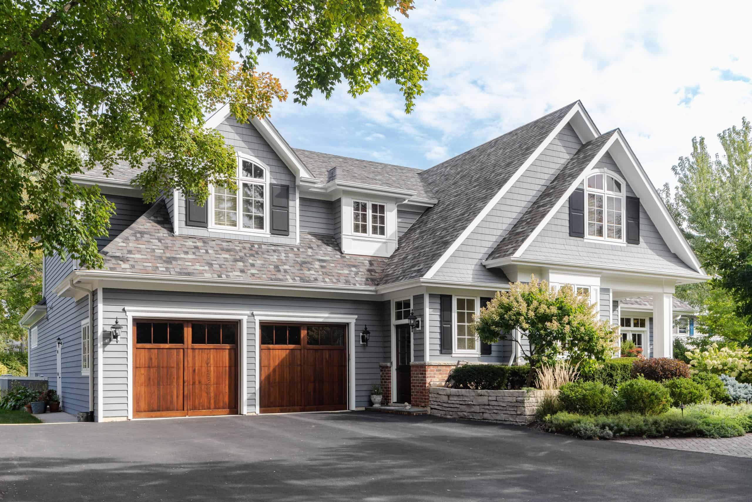 A two-story suburban home with gray siding and a gable roof. Recently enhanced by expert roofers, it features two wooden garage doors and a well-maintained garden. The driveway is wide, and mature trees surround the property. The sky is partly cloudy.