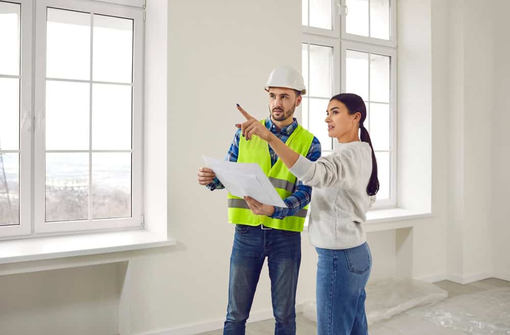 A woman and a man in a hard hat and safety vest stand in a bright room with large windows, discussing building plans. The woman points towards the windows while holding a blueprint.