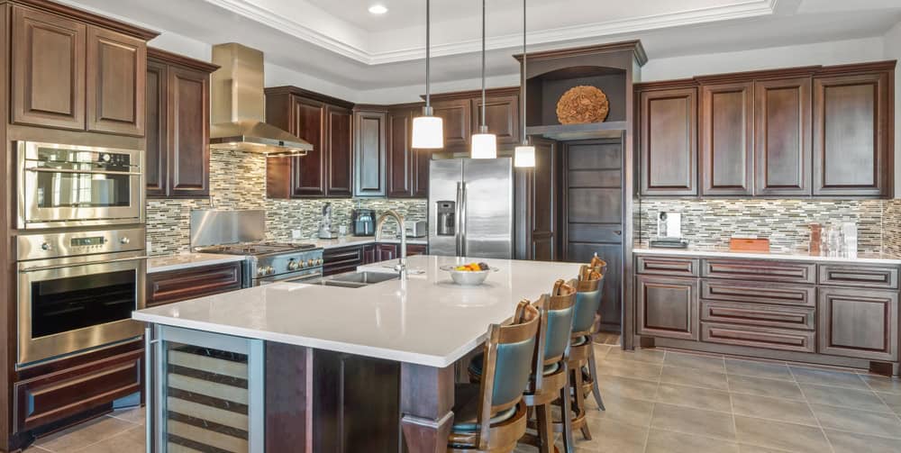 Spacious modern kitchen with dark wood cabinets, a large center island, and built-in stainless steel appliances. There are pendant lights hanging above the island, and the backsplash features a mosaic design. Four chairs line the island.