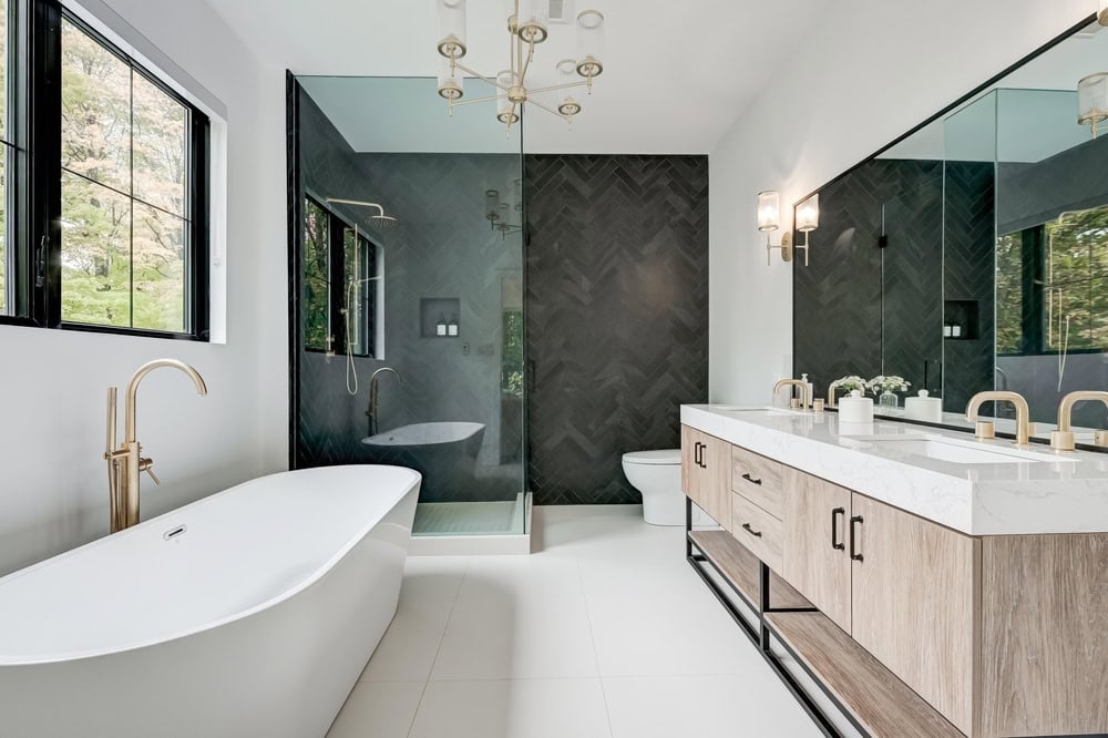 Modern bathroom with a freestanding bathtub, large glass-enclosed shower, and a double-sink vanity with marble countertops. The space features black herringbone wall tiles and large mirrors, with views of greenery through a window.