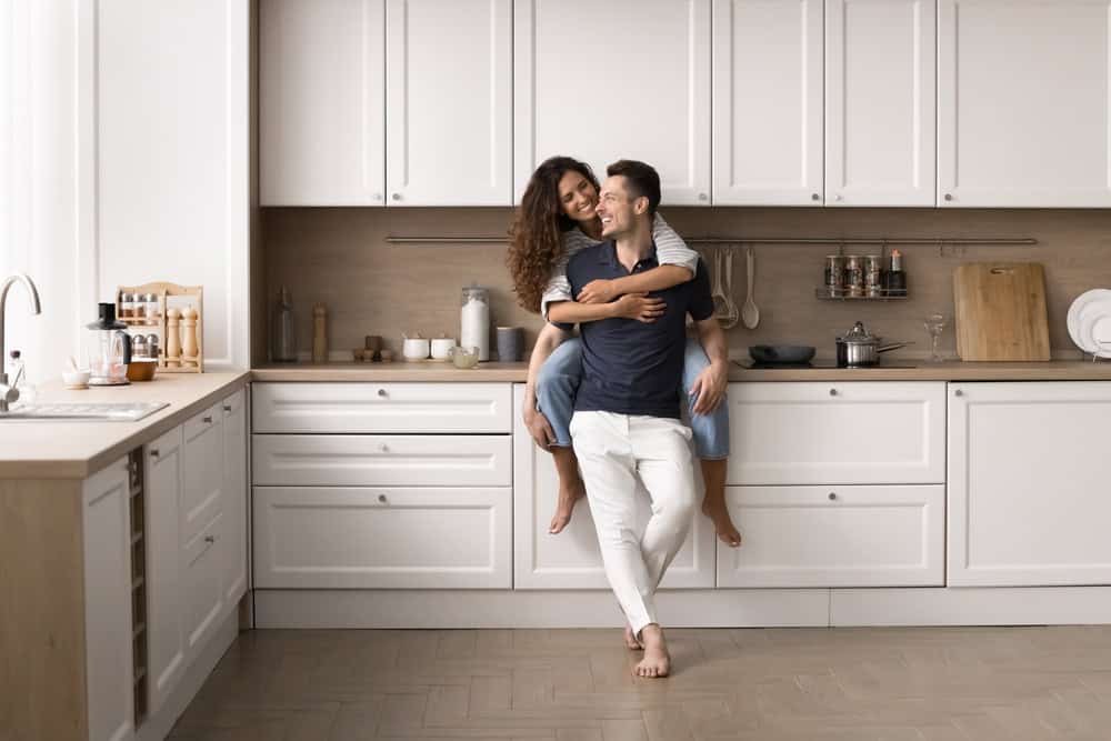 A couple is happily embracing in a modern kitchen with white cabinets and wooden countertops. The woman sits on a counter, hugging the man who stands barefoot, smiling. The kitchen has various utensils and containers neatly arranged.