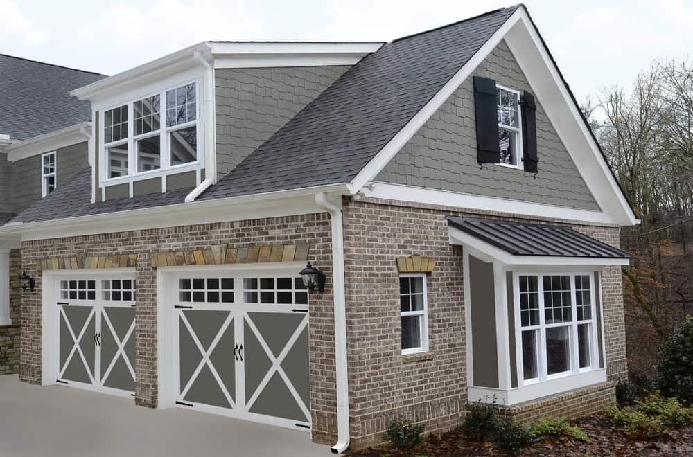 A suburban house with gray brick and siding. It features two large garage doors with a crossbeam design and a dormer window above. There are several windows and a sloped driveway. Trees are visible in the background.