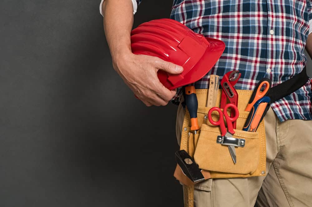 A person wearing a plaid shirt and beige pants is holding a red construction helmet. They have a tool belt around their waist containing scissors, a hammer, and other tools, standing against a dark background.