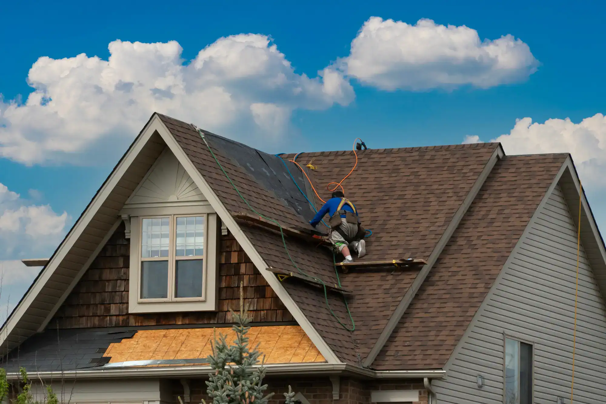 A worker wearing safety gear repairs the brown shingle roof of a house under a blue sky with clouds. Some shingles are missing, and tools and ropes are visible on the roof.