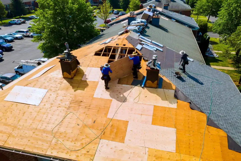 Three workers wearing safety gear repair a large sloped roof, replacing wooden panels and shingles. Tools and materials are scattered around, and trees and parked cars are visible in the background.