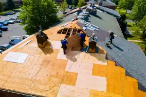 Three workers wearing safety gear repair a large sloped roof, replacing wooden panels and shingles. Tools and materials are scattered around, and trees and parked cars are visible in the background.