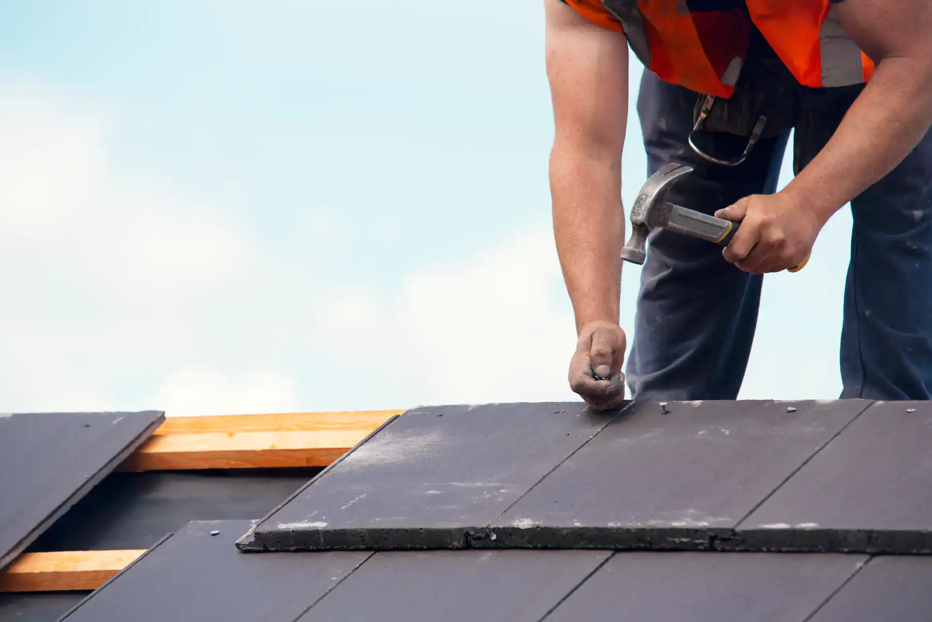 A construction worker wearing an orange safety vest uses a hammer to secure dark roof tiles, working on the sloped roof of a building under a clear sky.