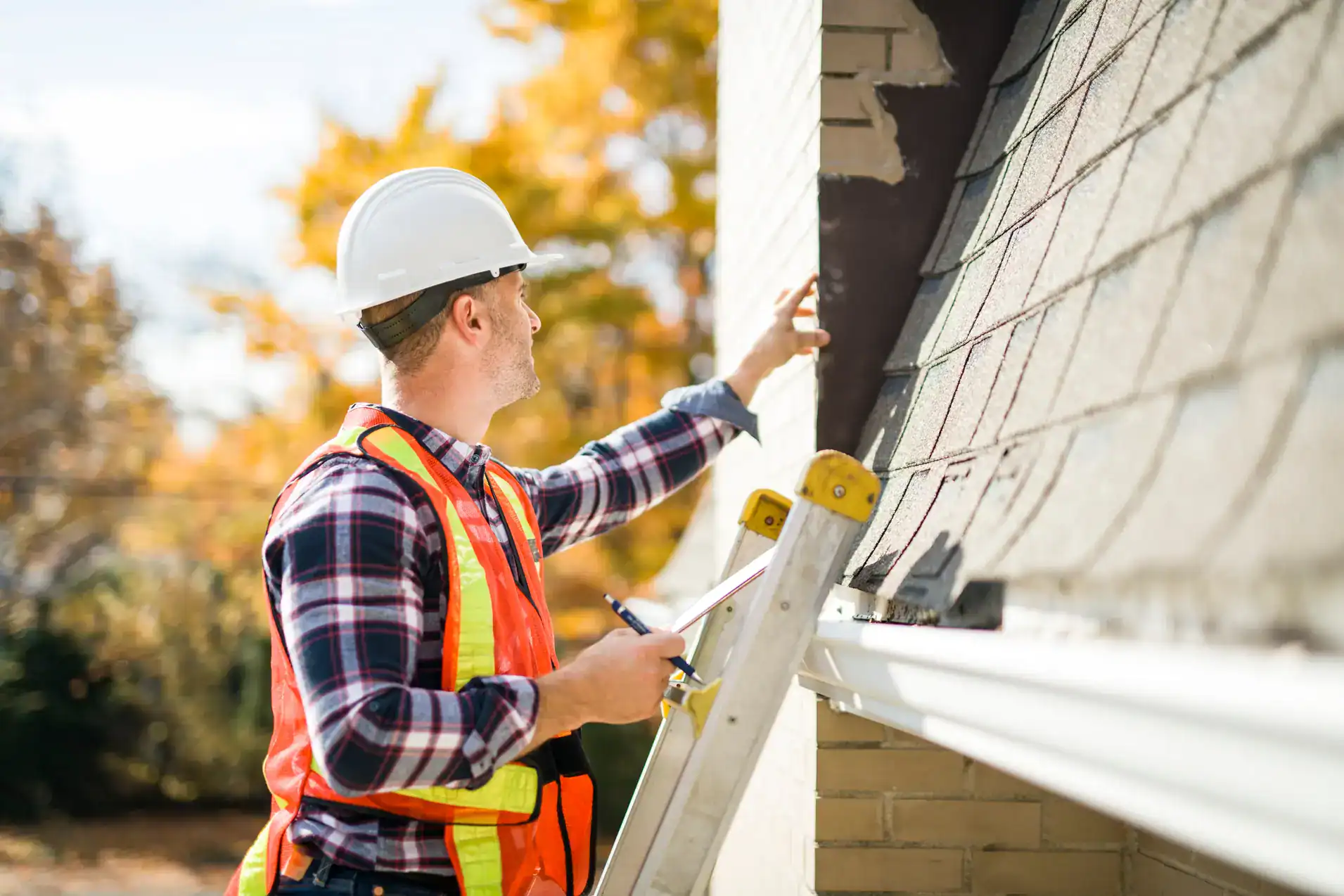A construction worker in a hard hat and reflective vest stands on a ladder, inspecting the exterior roof and siding of a house while holding a clipboard. Autumn trees are visible in the background.