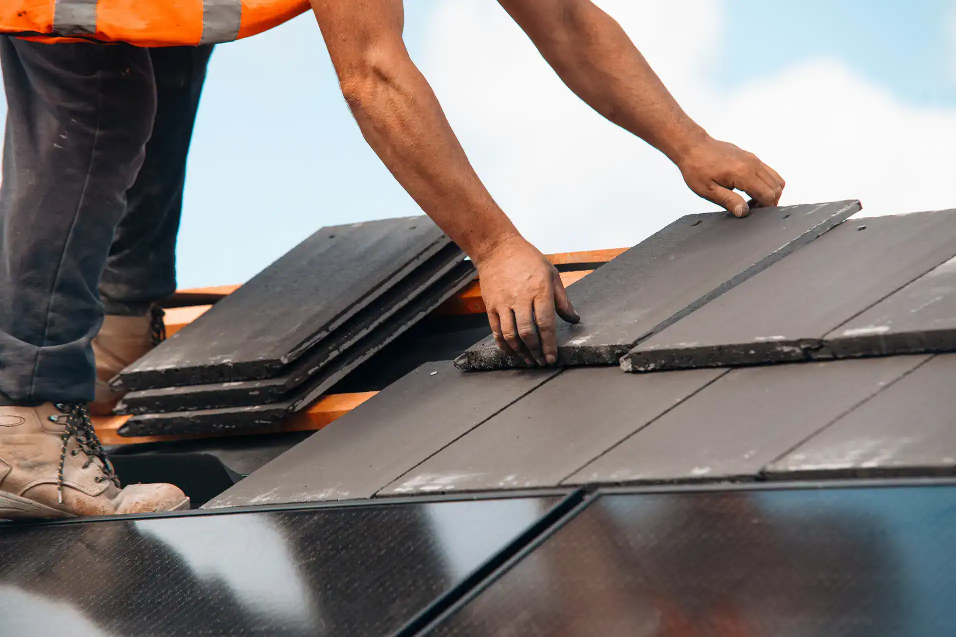A worker in boots and an orange vest installs dark roof tiles on a house under a blue sky. The worker is placing one tile while holding a stack of tiles with the other hand.
