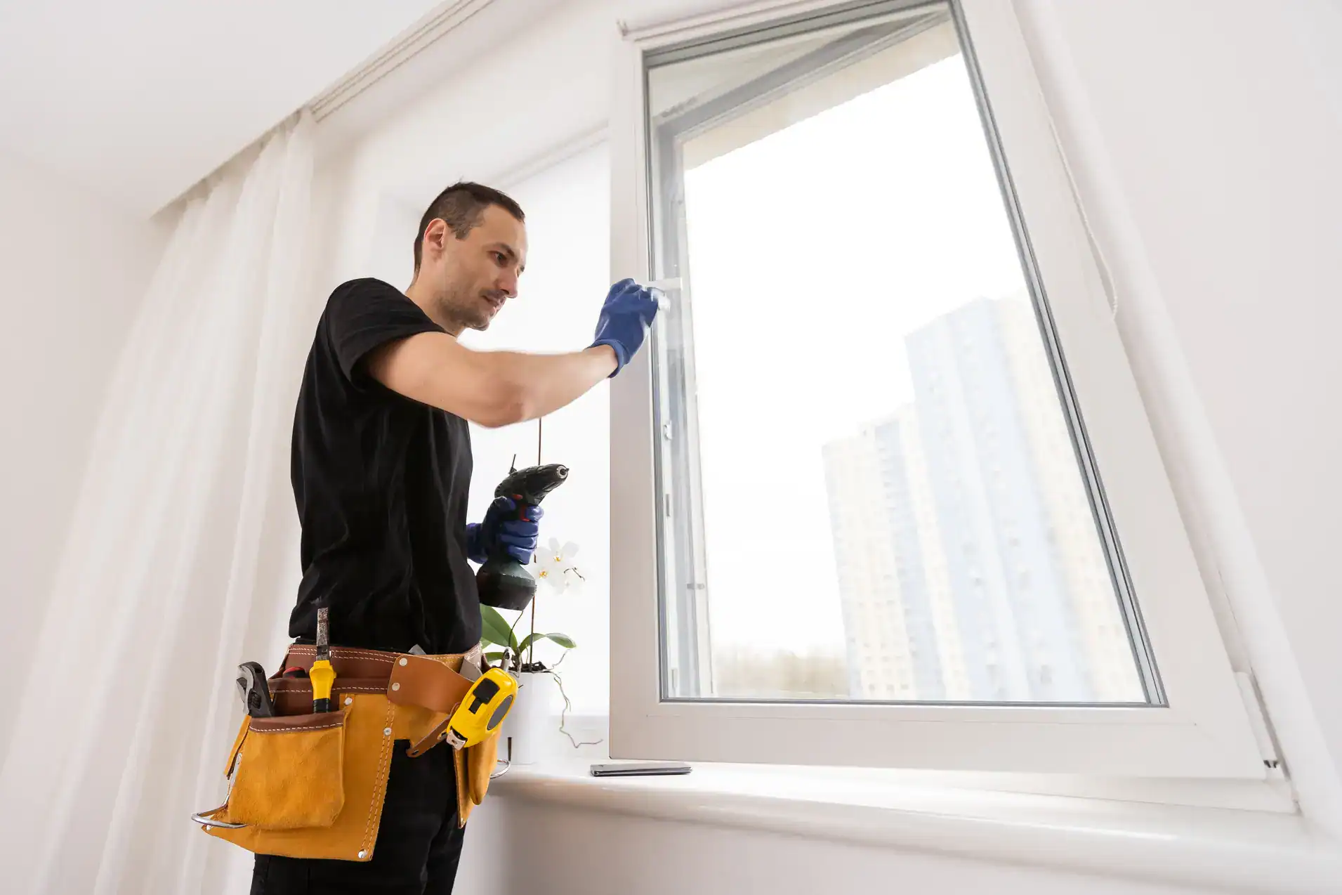 A man wearing gloves and a tool belt is working on an open window with a power drill in a bright room. A tall building is visible outside through the window.