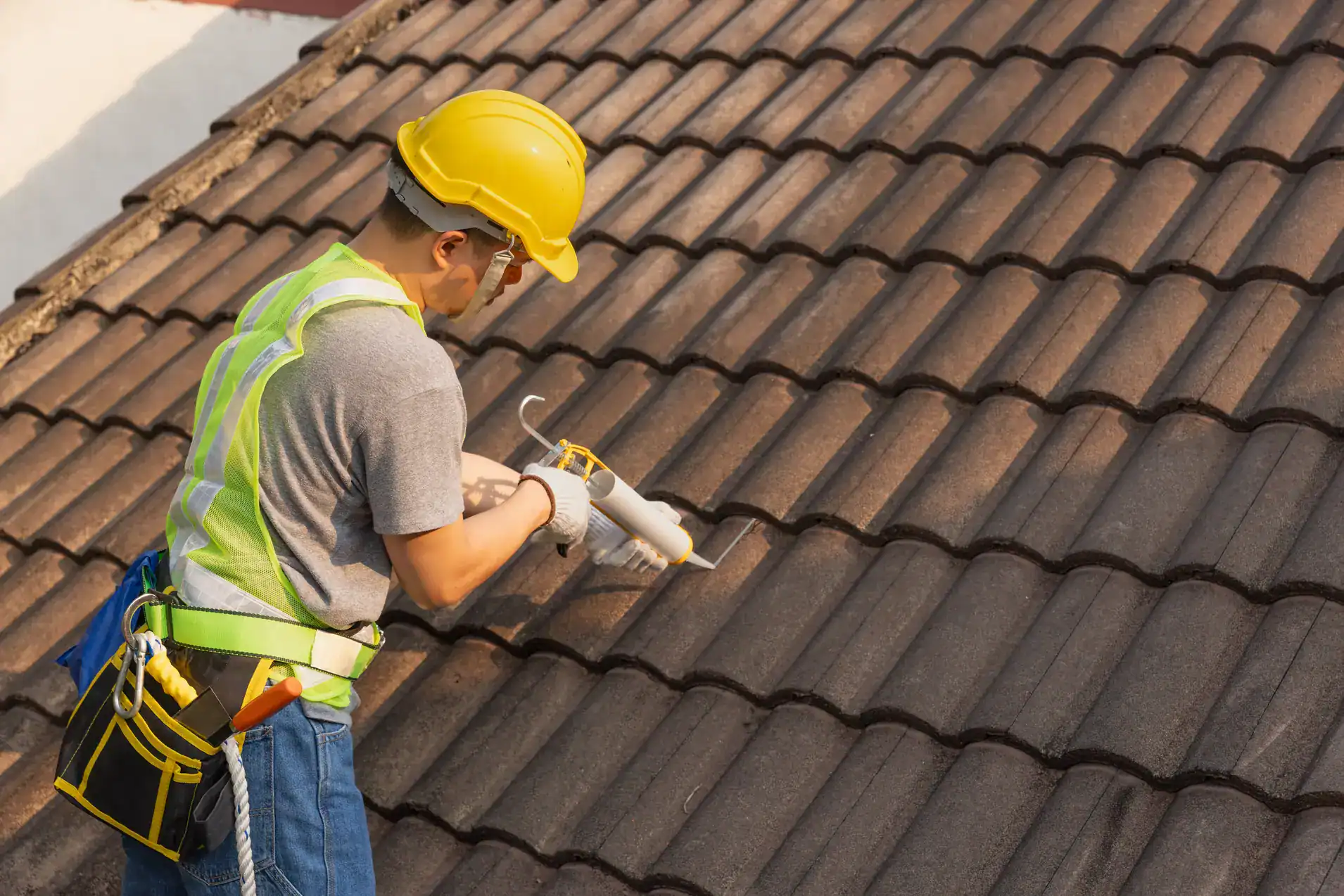 A construction worker wearing a yellow hard hat, safety vest, and tool belt applies sealant to the joints of brown roof tiles on a sloped roof during daytime.