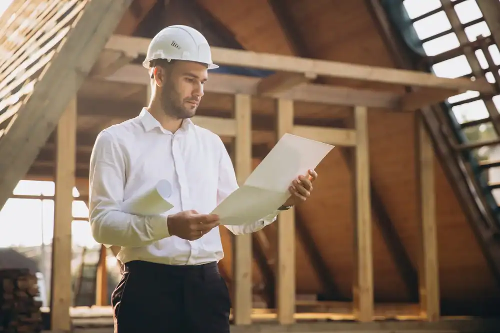 A man wearing a white shirt and a safety helmet, likely a general contractor Suffolk County, reviews architectural plans while standing at a NY construction site with a partially built wooden structure in the background.