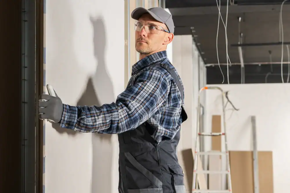 A construction worker wearing a plaid shirt, overalls, safety glasses, and a cap—typical of a general contractor Suffolk County, NY—uses a tool on a wall inside an unfinished building with exposed wiring and a ladder in the background.