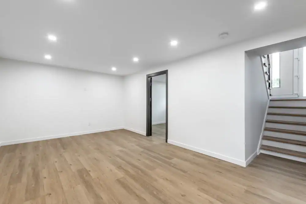 A bright, empty room with white walls, wood flooring, recessed ceiling lights, and a stairway leading up—expertly finished by a general contractor Suffolk County, NY—with a doorway opening to another inviting space.