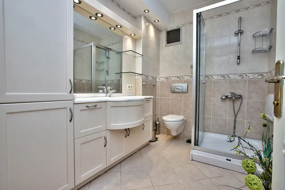 Modern bathroom in NY featuring white cabinets, large mirror, sink, wall-mounted toilet, glass shower enclosure, and beige tiled walls and floor. Upgrade your space with a trusted general contractor Suffolk County recommends.