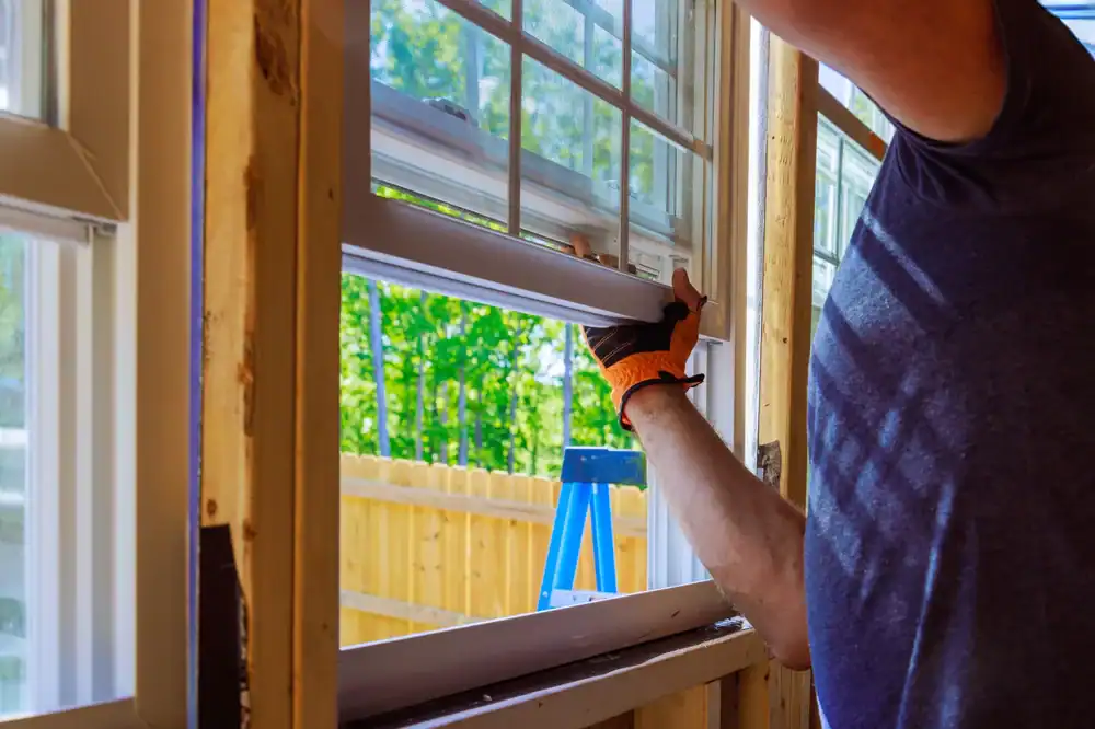 A person in gloves installs a window frame in a Suffolk County, NY home under construction with trees outside.